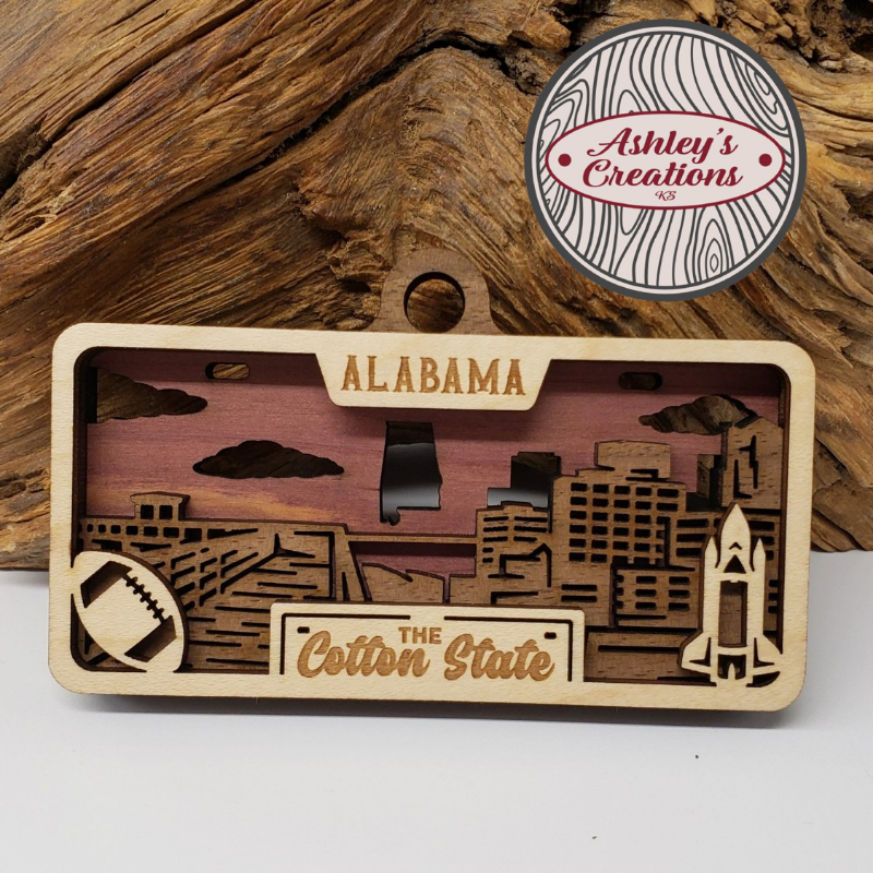 Wooden ornament with 'Alabama' and 'The Cotton State' text, featuring a cityscape design, on a wooden background.  Made by Ashley's Creations KS.