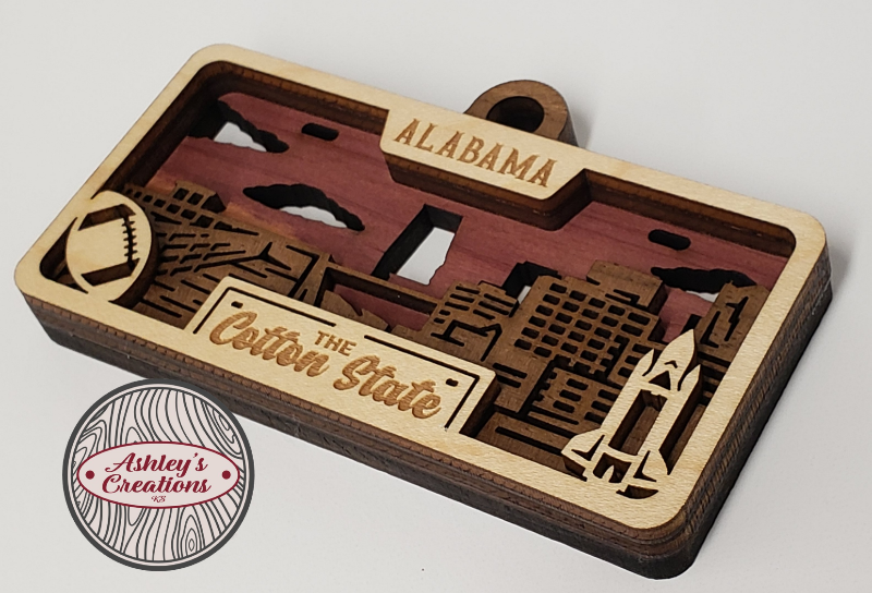 Wooden ornament with 'Alabama' and 'The Cotton State' text, featuring laser-cut shapes, on a light gray background.  Made by Ashley's Creations KS.