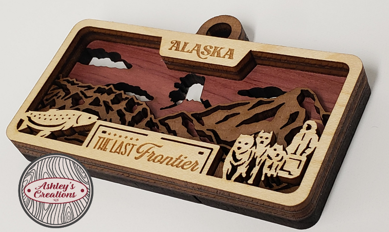 Wooden three layer license plate ornament with laser-cut designs and 'Alaska' text on a white background