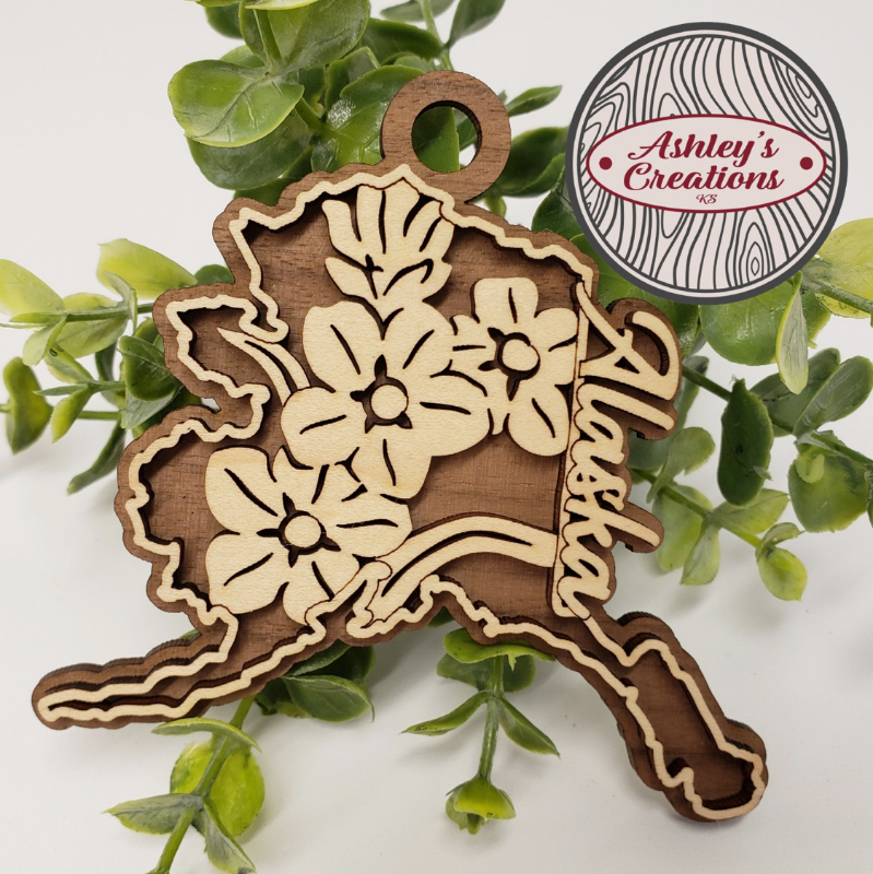 Wooden ornament shaped like the state of Alaska with floral designs on a green leaf background, featuring 'Ashley's Creations' logo.