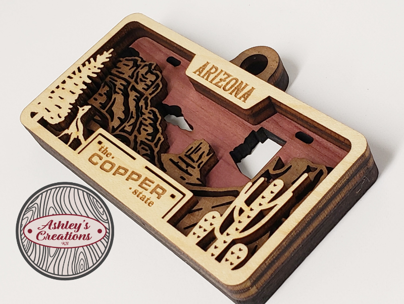 Wooden three layer license plate ornament with laser-cut designs and 'Arizona' text, featuring 'Ashley's Creations' logo.