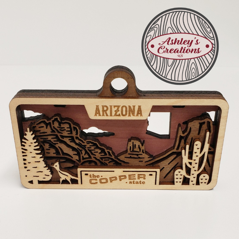 Wooden three layer license plate ornament of Arizona with desert landscape and 'The Copper State' text, branded 'Ashley's Creations'.