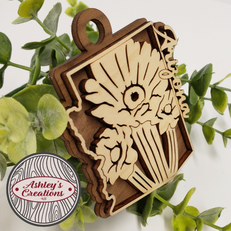 Wooden ornament with floral design and 'Ashley's Creations' logo on a white background with green leaves.