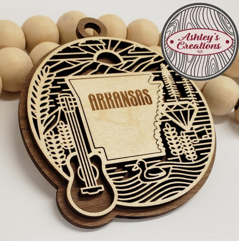 Wooden ornament with 'Arkansas' engraving, surrounded by wooden beads, branded 'Ashley's Creations'.