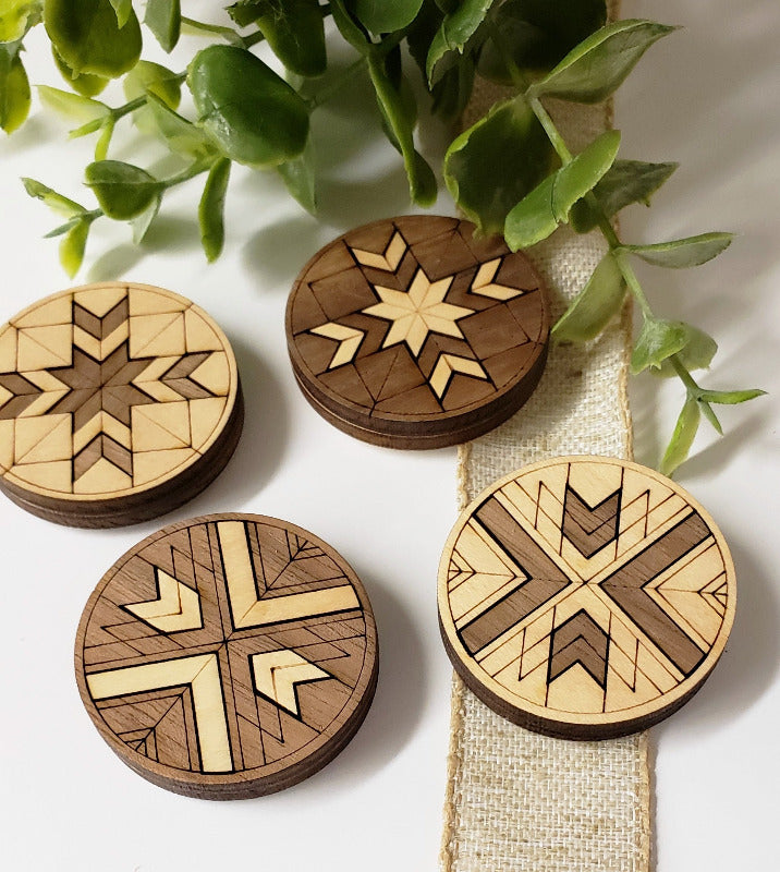 A set of four wooden magnets with geometric barn quilt patterns displayed on a white surface next to a green plant.
