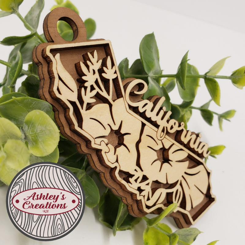 Wooden ornament shaped like the state of California with 'California' engraved, surrounded by green leaves.