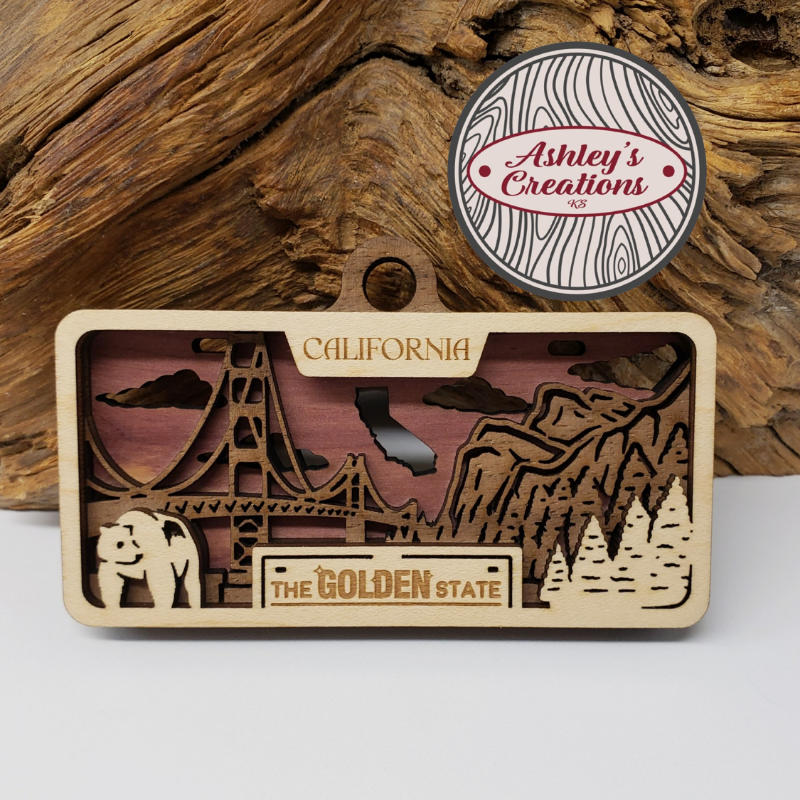 Wooden three layer license plate ornament with laser-cut designs and 'Ashley's Creations' logo on a wooden background