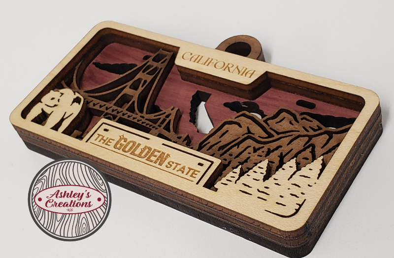 Wooden three layer license plate ornament with laser-cut designs of a ship, mountains, and text on a white background.
