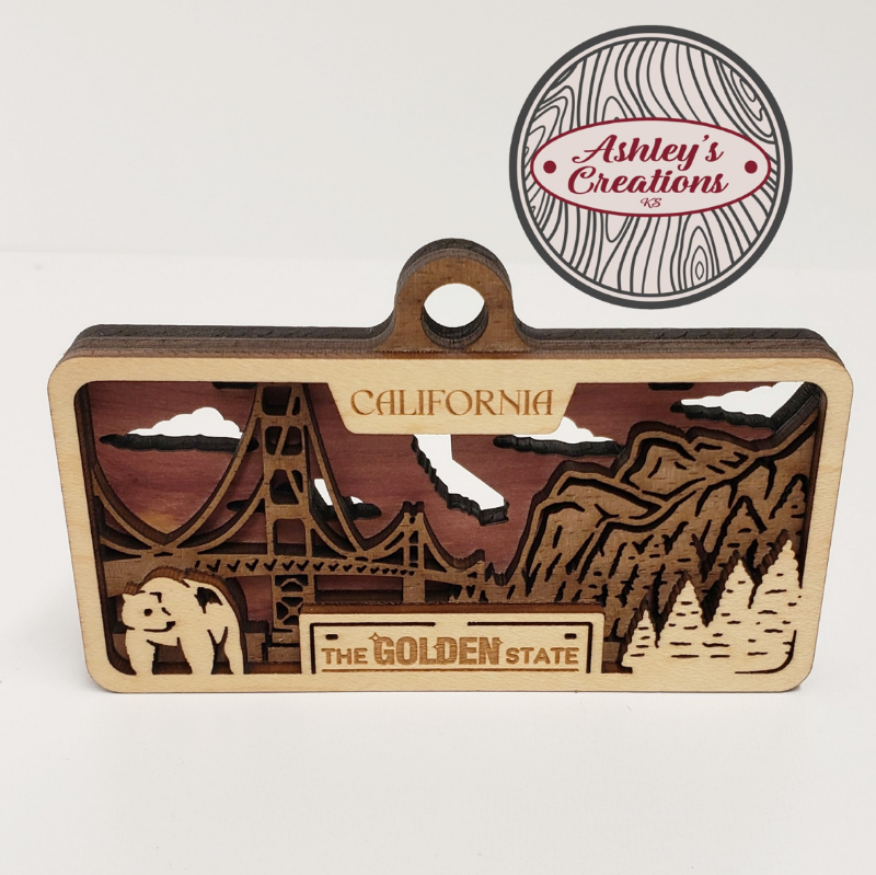 Wooden three layer license plate ornament with laser-cut design of a bridge and mountains, featuring 'Ashley's Creations' logo.