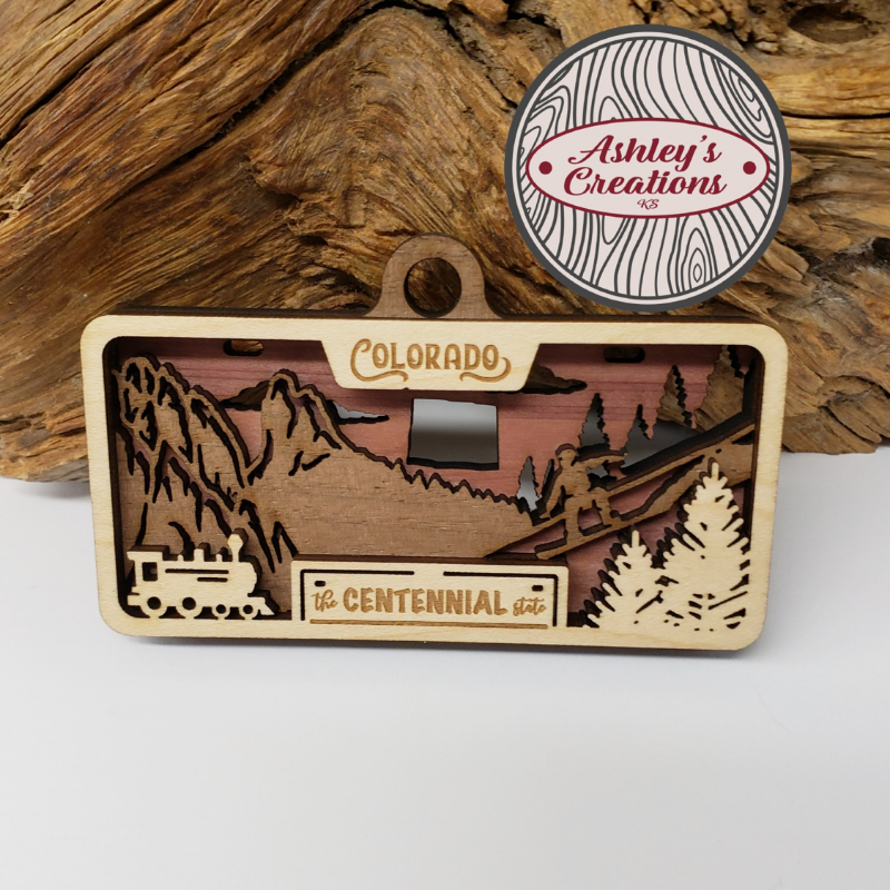 Wooden three layer license plate ornament with mountain and train design, featuring 'Ashley's Creations' logo.