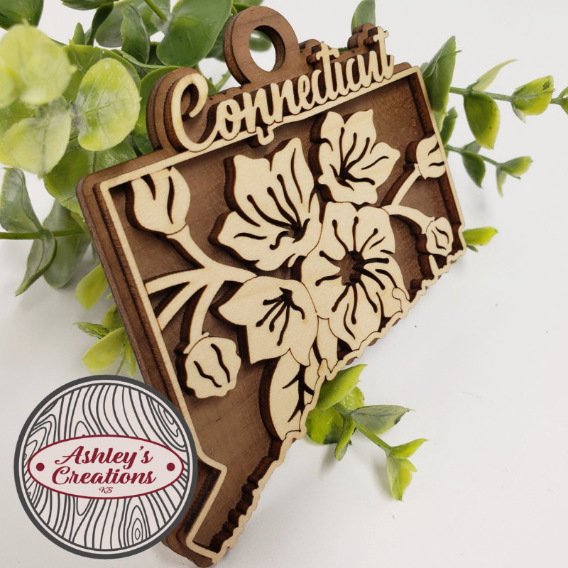 Wooden ornament shaped like the state of Connecticut with floral design and 'Connecticut' text, surrounded by green leaves.