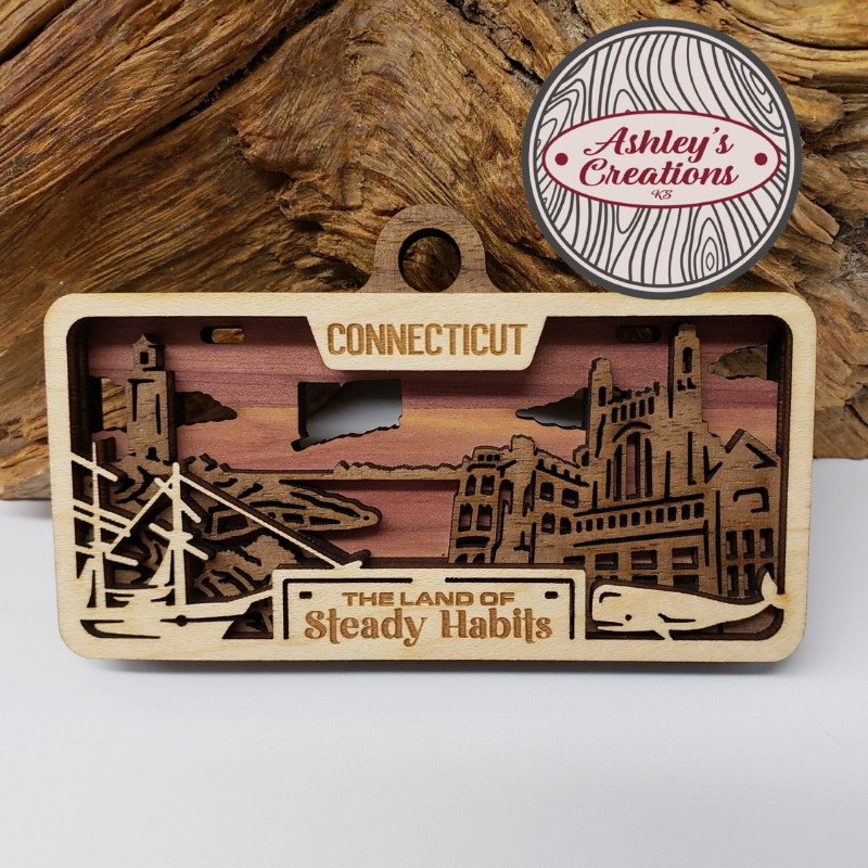 Wooden three layer license plate ornament with laser-cut designs and text, branded 'Ashley's Creations', on a wooden background.