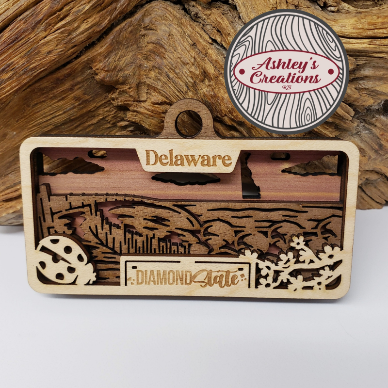 Wooden three layer license plate ornament with 'Delaware' and 'Diamond State' text, featuring a small round badge with 'Ashley's Creations' on a wooden background.