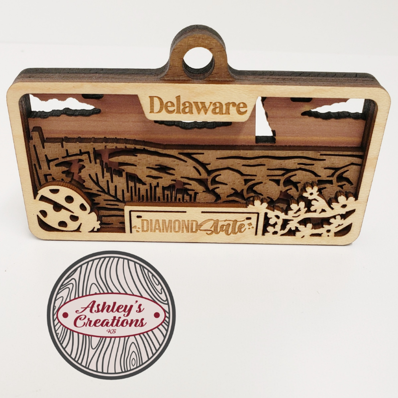 Wooden three layer license plate ornament with laser-cut designs and 'Delaware' text, featuring 'Ashley's Creations' logo.
