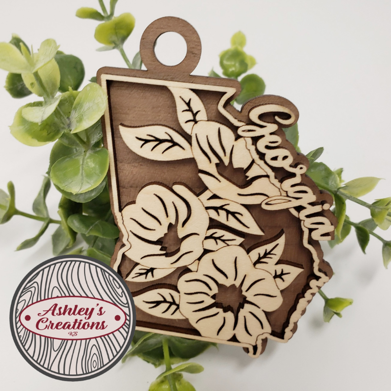 Wooden ornament with floral patterns and 'Cheer' text, surrounded by green leaves, with 'Ashley's Creations' logo.