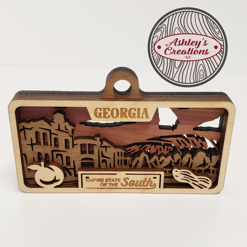Wooden three layer license plate ornament with laser-cut designs and 'Georgia' text, branded 'Ashley's Creations'.