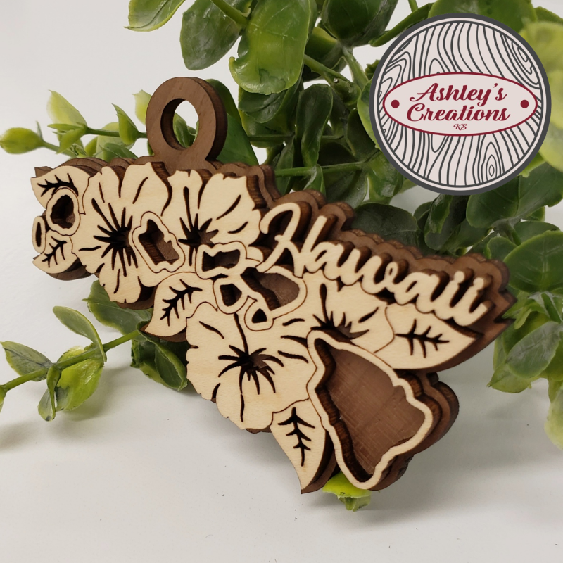 Wooden ornament shaped like Hawaii with floral designs on a green leaf background, branded 'Ashley's Creations'.