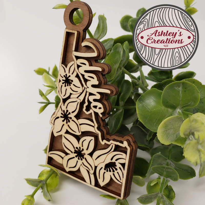 Wooden ornament with floral patterns on a green leaf background, featuring 'Ashley's Creations' logo.