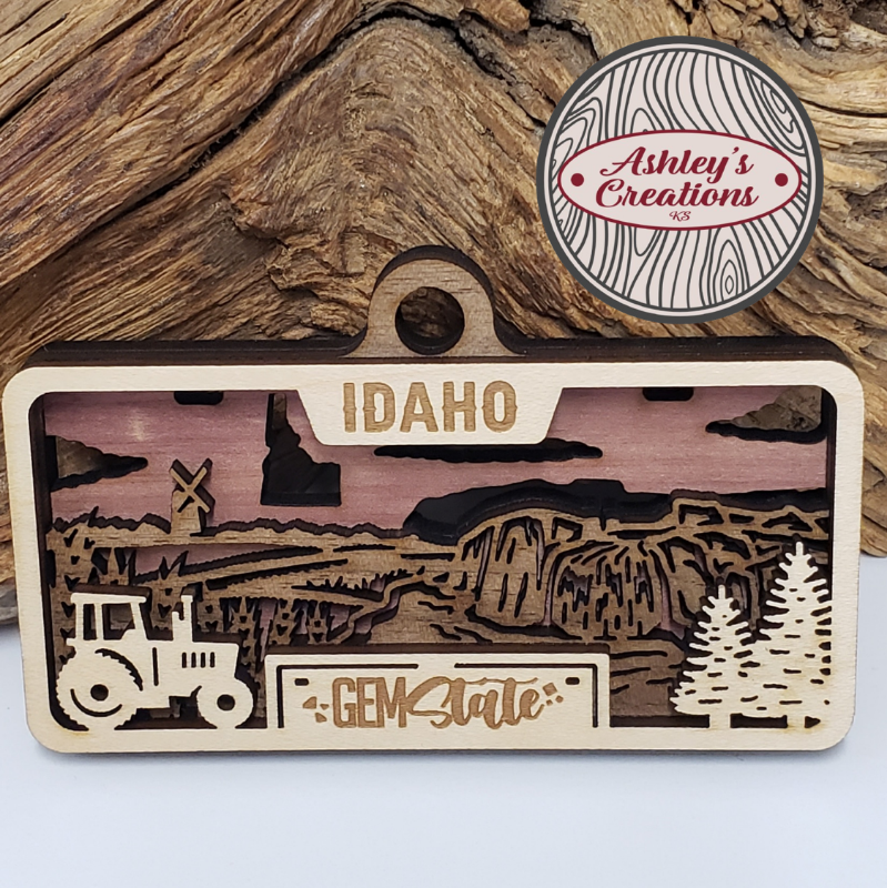 Wooden Idaho-themed three layer license plate ornament with laser-cut landscape and text, branded 'Ashley's Creations'.