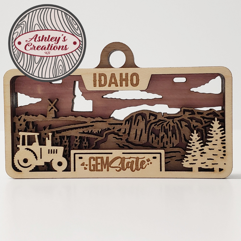 Wooden three layer license plate ornament with Idaho landscape and 'Gem State' text, featuring a tractor and trees.