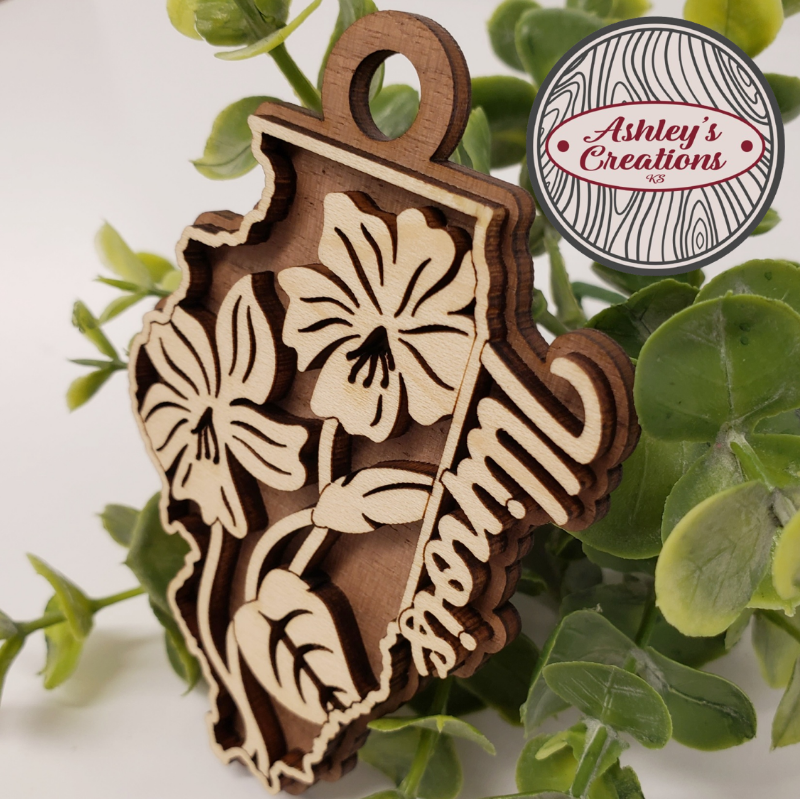 Wooden ornament with floral design on a green leaf background, featuring 'Ashley's Creations' logo.