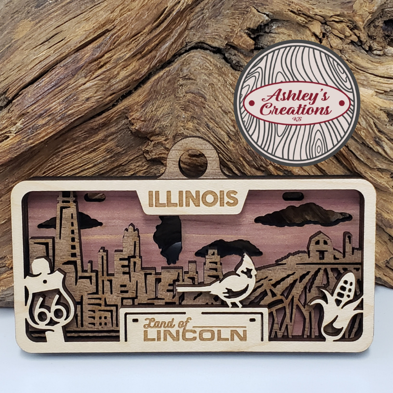 Wooden three layer license plate ornament with 'Illinois' and 'Land of Lincoln' text, featuring a bird and corn stalk design, on a wooden background.