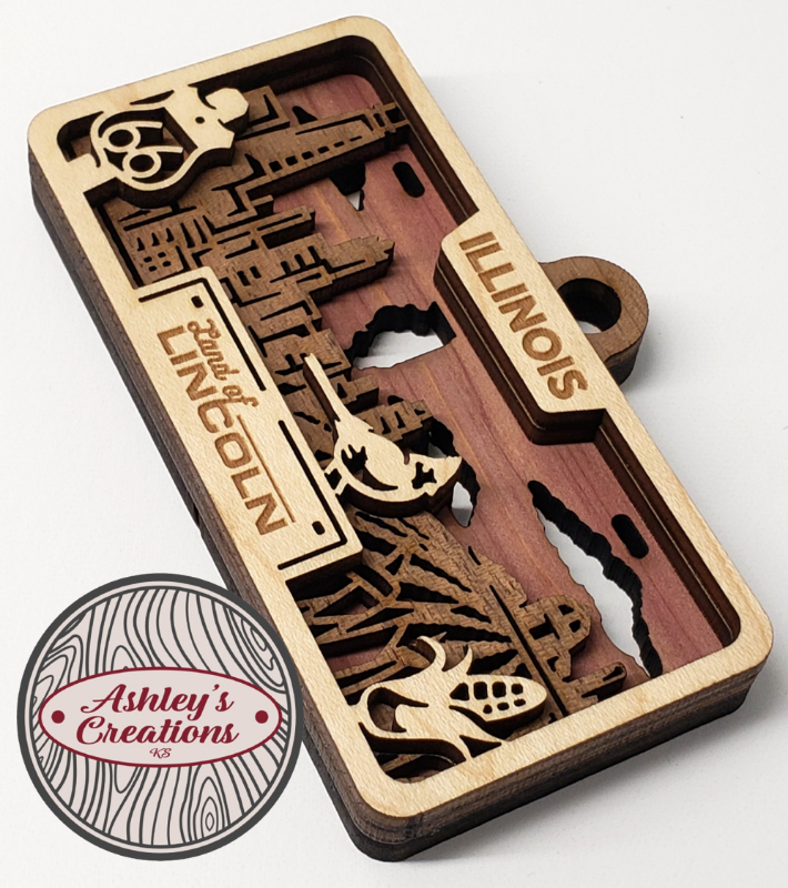 Wooden three layer license plate ornament with intricate cut-out designs on a white background, featuring the brand 'Ashley's Creations'.