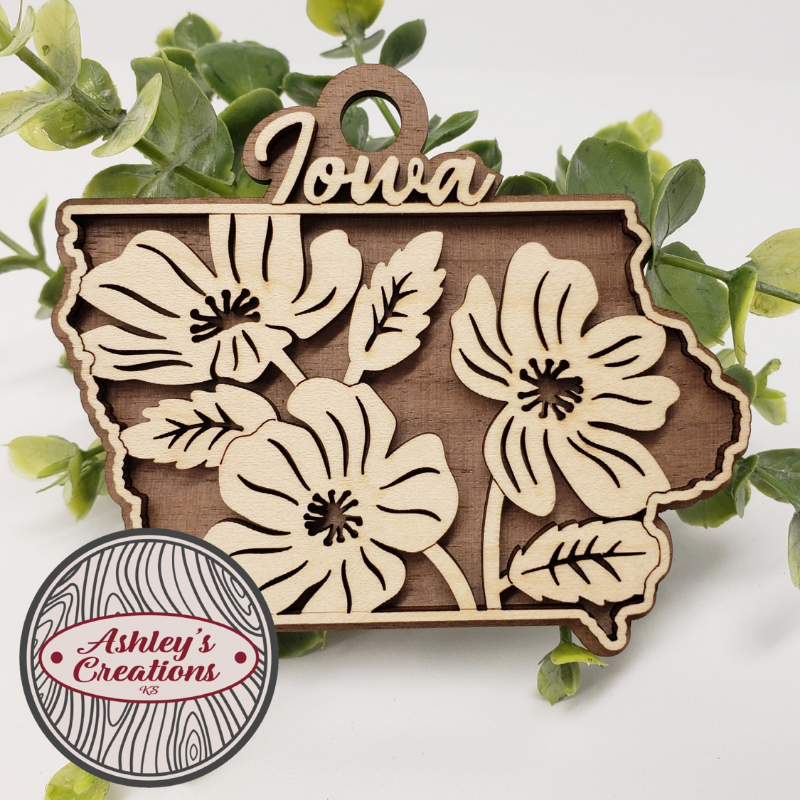Wooden ornament shapeed like the state of Iowa with floral designs and 'Iowa' text, surrounded by green leaves.