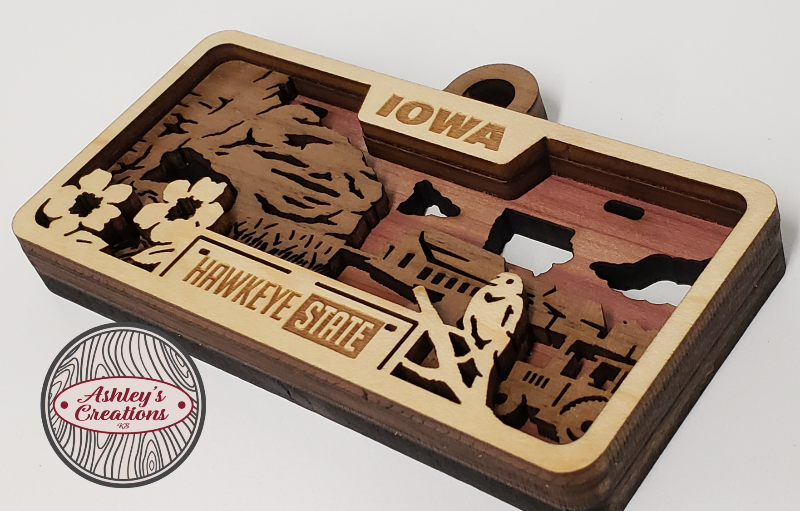 Wooden ornament with laser-cut designs and 'Iowa Hawkeye State' text, featuring a logo in the corner.