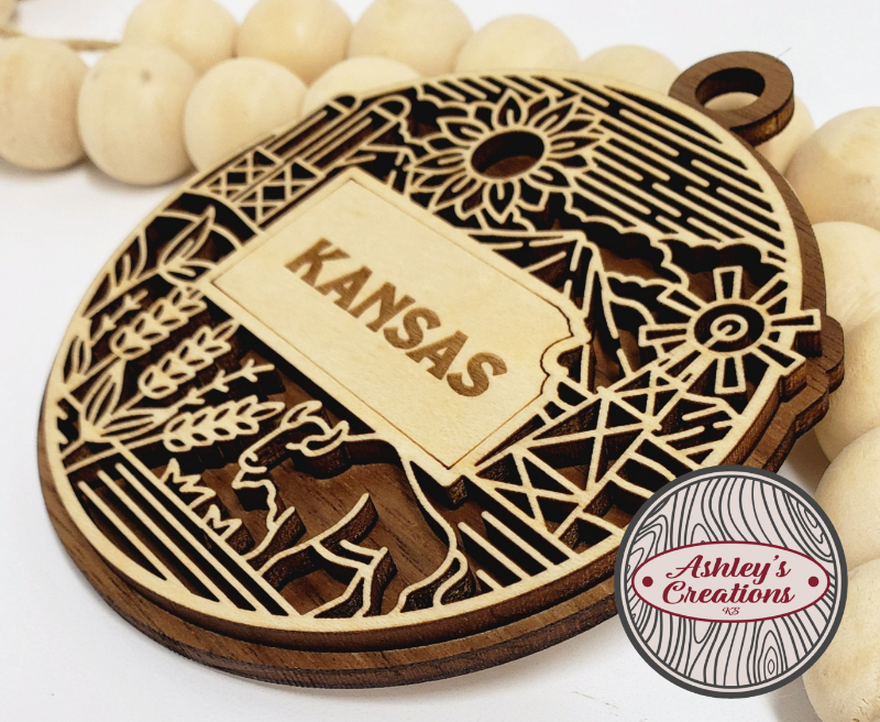Wooden ornament with 'Kansas' engraving surrounded by wooden beads, branded 'Ashley's Creations'.