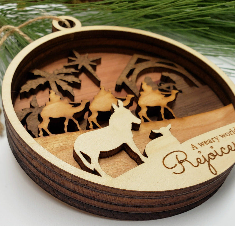 Wooden nativity scene ornament with cut-out figures and 'Rejoice' text, set against a natural background.
