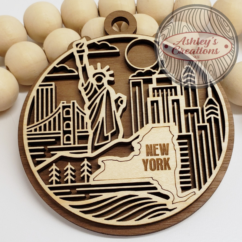 Wooden ornament with laser-cut design of the Statue of Liberty and New York skyline, branded 'Ashley's Creations'.