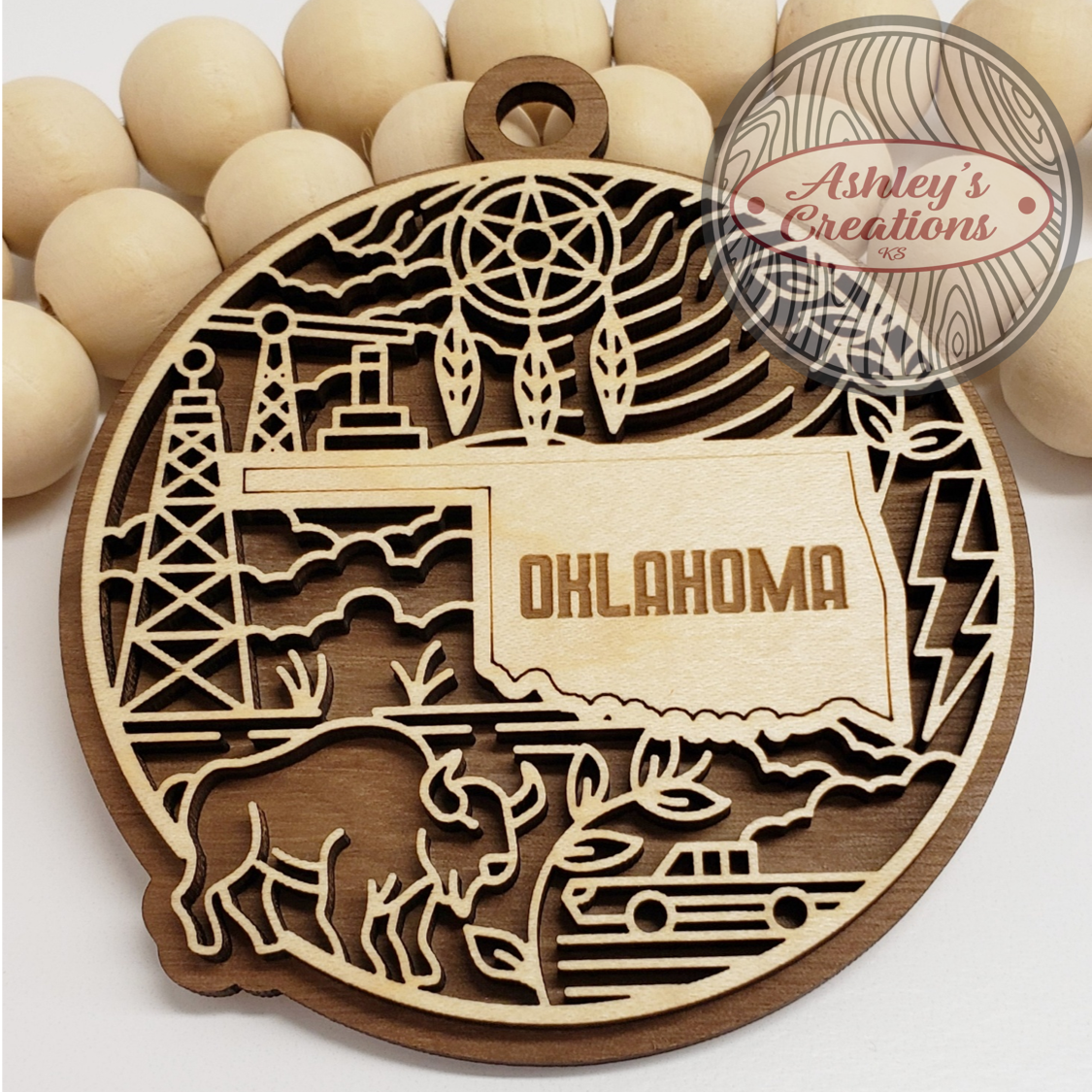 Wooden ornament with laser-cut designs of Oklahoma, a deer, and a truck, branded 'Ashley's Creations'.