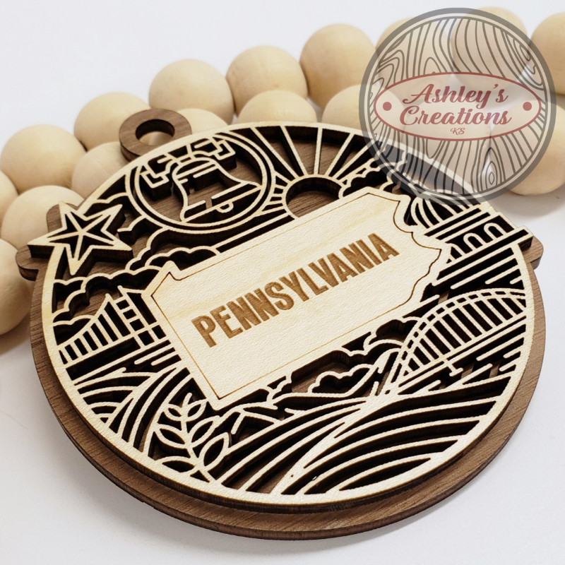 Wooden ornament with 'Pennsylvania' text and bell design, branded 'Ashley's Creations'.